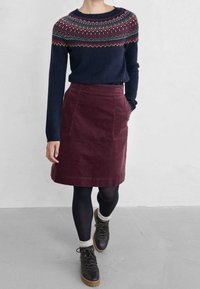 Pull bleu marine avec un empiècement à motifs en rouge et vert, associé à une jupe en velours côtelé bordeaux. Collants noirs et bottines à lacets avec des accents crème.