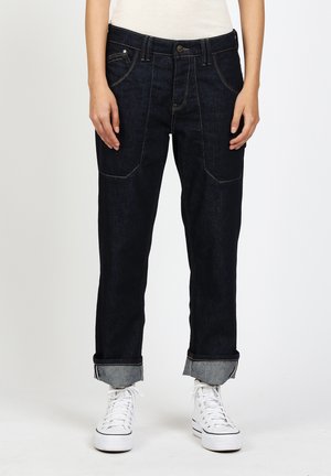 Person wearing dark denim rolled-up jeans and white high-top sneakers, standing against a plain light background.