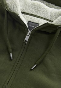 Olivengrøn zip-up hoodie med en blød cremefarvet sherpa-foring, metalzipper og snore. Har et label med teksten "NEXT" indeni.