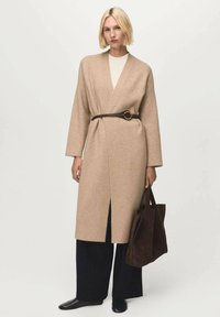 Donna bionda che indossa un cardigan beige con cintura sopra una maglietta bianca, pantaloni neri a gamba larga, scarpe nere e tiene in mano una grande borsa a tracolla in suede marrone.