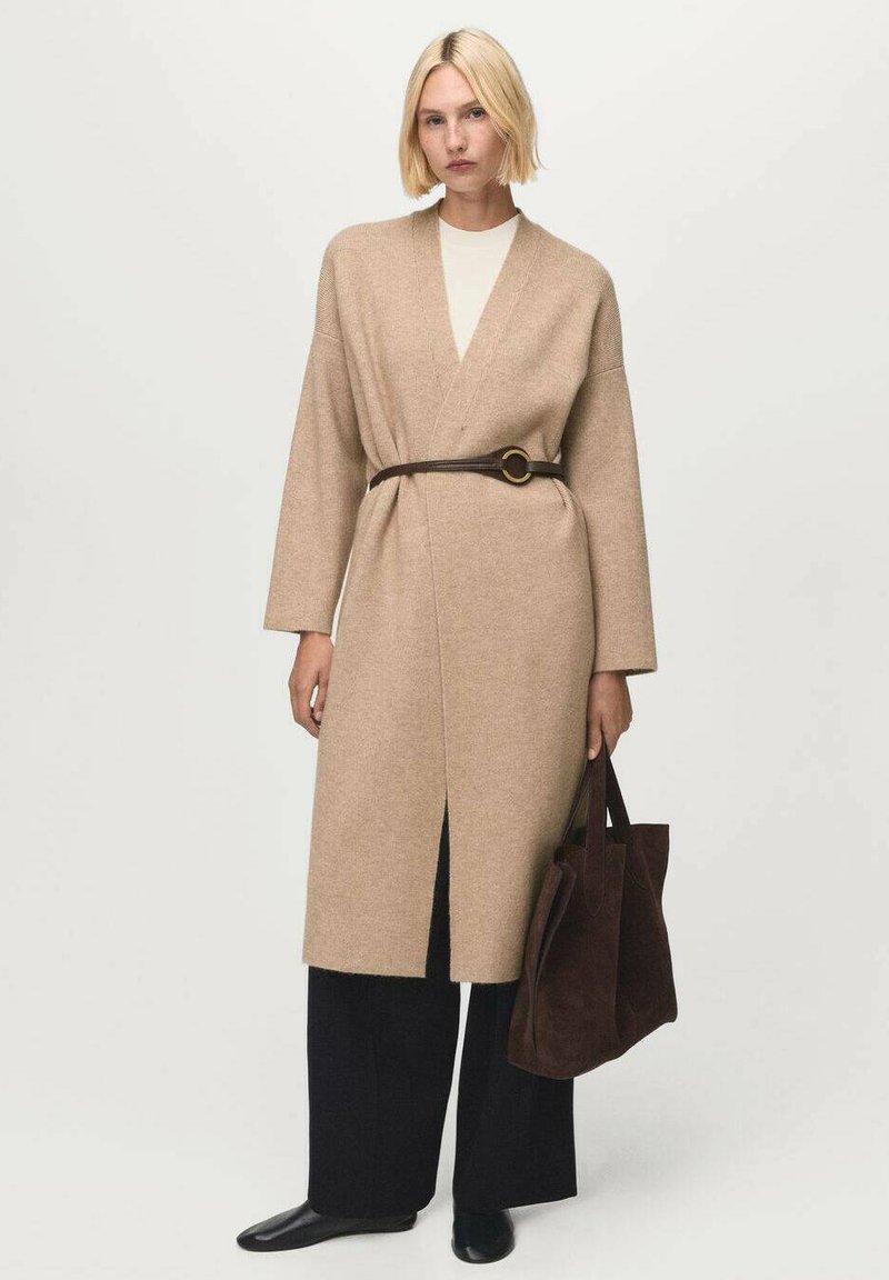 Donna bionda che indossa un cardigan beige con cintura sopra una maglietta bianca, pantaloni neri a gamba larga, scarpe nere e tiene in mano una grande borsa a tracolla in suede marrone.