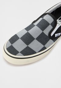 Chaussure à enfiler avec motif damier noir et gris, semelle en caoutchouc blanche et bordure noire, marquée avec une étiquette Vans.