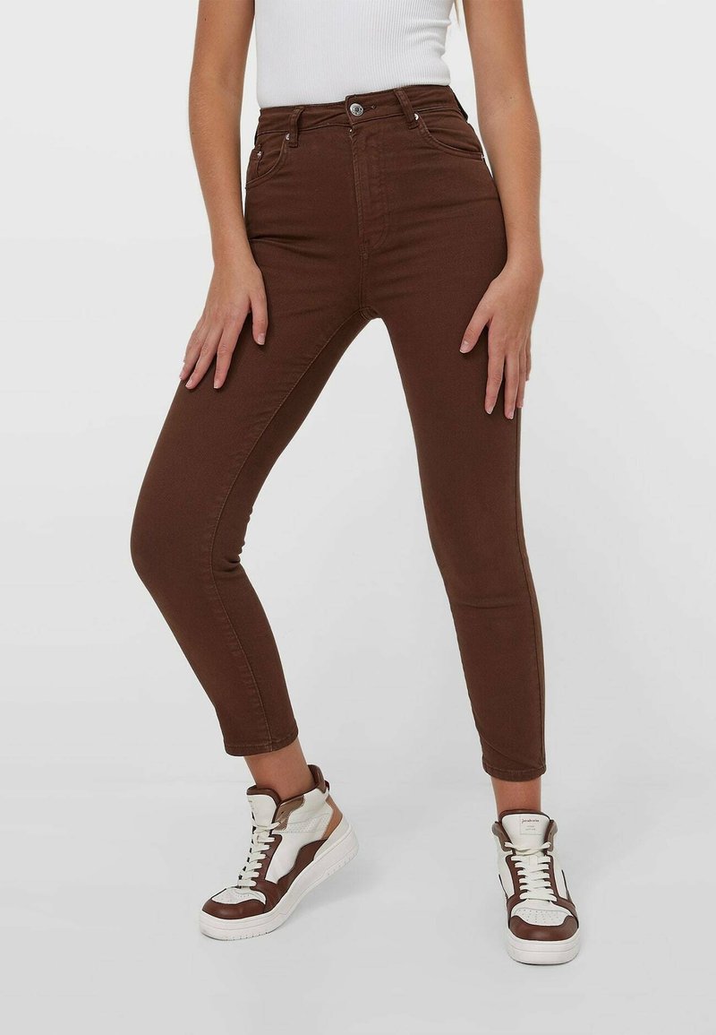 Femme portant un jean skinny marron taille haute et des baskets montantes blanches et marron, debout devant un fond blanc uni.