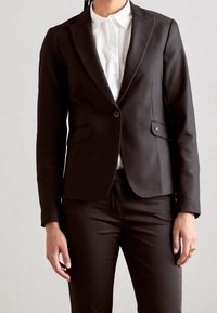 Blazer negro de corte entallado, con solapas de muesca y dos bolsillos delanteros. Se lleva sobre una camisa blanca con cuello, combinada con pantalones negros ajustados.