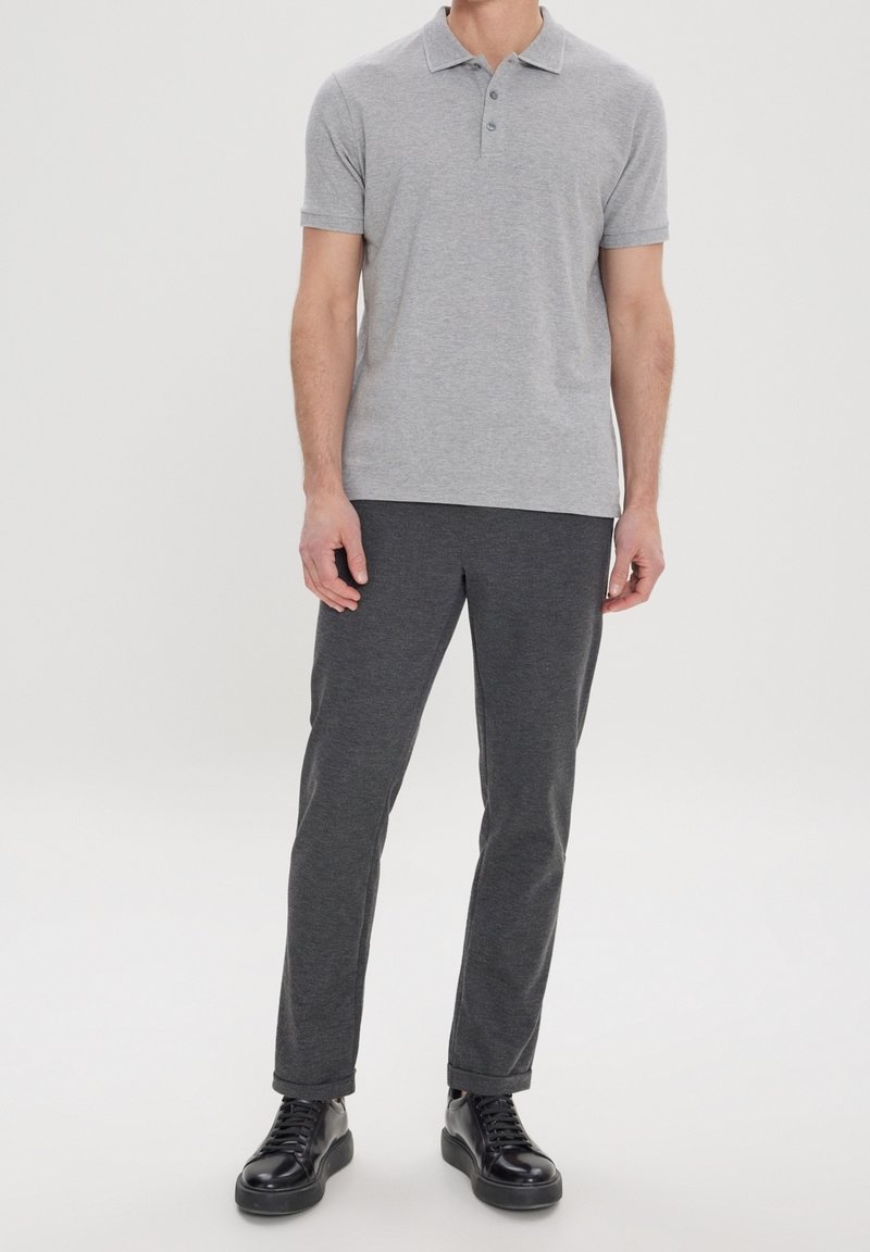 Homme portant un polo à manches courtes gris clair, un pantalon gris foncé et des baskets en cuir noir, debout devant un fond blanc uni.