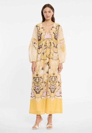 Frau trägt ein langes, fließendes Kleid mit weiten Ärmeln und einem gelben, rosa und braunen, kunstvollen Blumen- und Paisley-Muster, kombiniert mit weißen Sandalen mit Absatz.