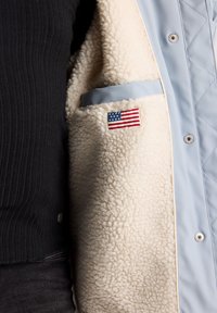 Valkoinen vuorillinen vaaleansininen takki, jossa on kermaisenvalkoinen turkisvuori. Takissa on ulkopuolella tasku, jossa on pieni amerikkalainen lippumarssi.