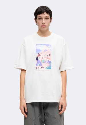 Persoon met kort donker haar die een wit T-shirt draagt met een pastelgekleurde wandelafbeelding en een grijze broek, staand tegen een effen achtergrond.