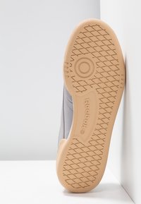 Graue Sneaker mit einer strukturierten, beige-gummiartigen Sohle, ausgestattet mit einem geometrischen Profilmuster und dem "Reebok"-Logo an der Seite. Minimalistisches Design.