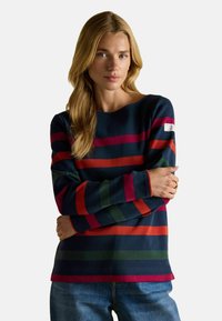 Žena s dlhými blond vlasmi, oblečená v tmavomodrom svetri s červenými, zelenými a ružovými vodorovnými pruhmi a modrými džínami, stojaca s prekríženými rukami.