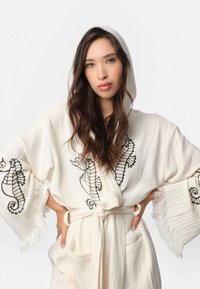 Biela róba s kapucňou, zdobená čiernym výšivkou morskej koníka, s frgňami na rukávoch a viazaním v páse. Obsahuje dve predné vrecká.