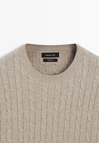 Béžový pletený bavlněný svetr s kulatým výstřihem, zdobený vzorem copánkového pletení a žebrovanými manžetami. Štítek viditelný uvnitř límce.
