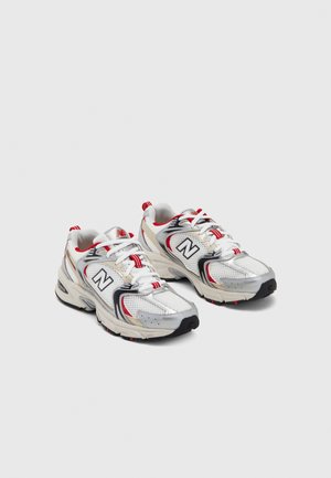 Chaussures de sport New Balance blanches, argentées et rouges avec un design en mesh, lacets et détails noirs, montrées côte à côte sur un fond uni.