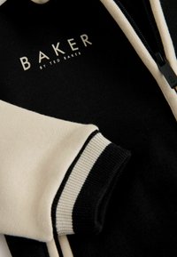 Sort og cremefarvet lynlåsbeklædning med ribbede kanter. Har et logo med teksten "BAKER by TED BAKER" i hvid. Blødt stof, sporty design.