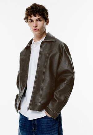 Brown faux leather jacket with a collared neckline, horizontal seam details, and zip closure, paired with a white shirt and blue jeans.