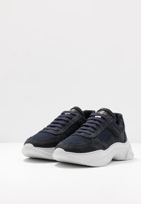 Sneaker blu scuro con pannelli in rete e suede, suole bianche spesse e lacci blu, posizionati affianco su una superficie bianca.