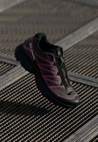 Black trail running shoe with pink accents, mesh upper, textured outsole, and a secure lacing system. Features a structured design.