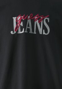 Camiseta negra con la palabra "guess" bordada en rojo y "JEANS" en letras plateadas agrietadas. Textura suave sin patrones adicionales.