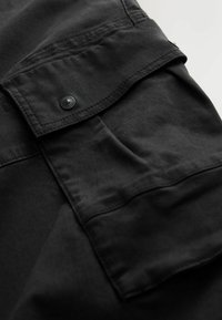 Black fabric, featuring a flap pocket with a black snap button. Smooth texture with subtle stitching details, emphasizing minimalist design.