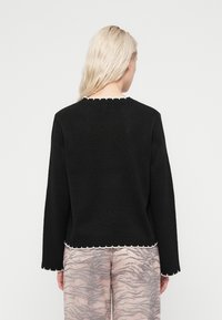 Μαύρο πλεκτό πουλόβερ με λευκή διακοσμητική φάσα με κυματοειδή όψη στον λαιμό και το τελείωμα, με μακριά μανίκια και άνετη εφαρμογή, συνδυασμένο με ανοιχτόχρωμο παντελόνι με σχέδια.