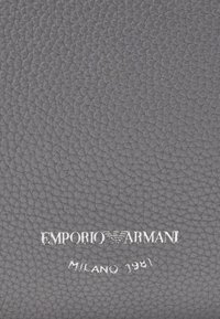 Harmaa nahkatekstuuri, jossa on rakeinen pinta. Siinä on hopeinen "Emporio Armani" -logo ja "Milano 1981" kaarevassa muotoilussa.