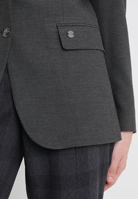Blazer gris foncé sur mesure avec poche à rabat et bouton en métal, porté avec un pantalon à carreaux foncé, une main visible sur le côté.