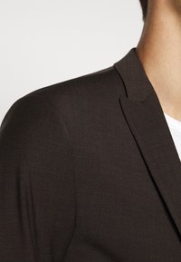 Hnědý blazer s texturovanou tkaninou. Má klopový límec a přiléhavý střih, zobrazený od ramene až k horní části hrudníku.