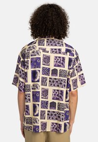 Short-sleeve shirt with a cream background featuring a collage of purple patterned squares, showcasing various floral and geometric designs.