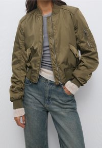 Persona con una chaqueta bomber verde olivo sobre una camiseta gris, con puños de punto blancos y jeans de tiro alto azules, con una mano en el bolsillo.