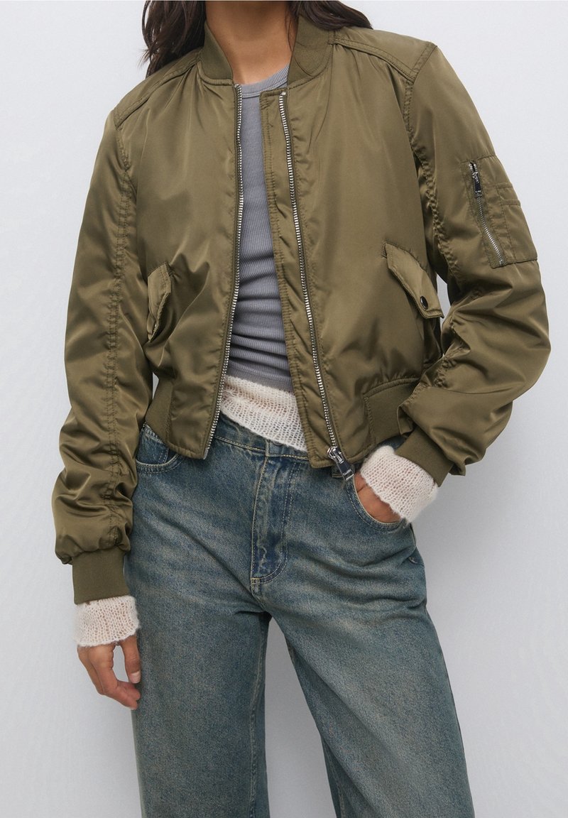 Persona con una chaqueta bomber verde olivo sobre una camiseta gris, con puños de punto blancos y jeans de tiro alto azules, con una mano en el bolsillo.