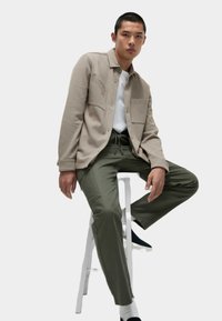 Veste en coton beige clair avec deux poches avant, portée sur un t-shirt blanc, accompagnée de pantalons vert olive. Positionnée sur un tabouret blanc.