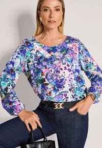 Bunte Bluse mit floralen Mustern in Lila, Blau und Pink. Lange Ärmel, runder Ausschnitt, kombiniert mit dunklen Jeans und einer schwarzen Handtasche.