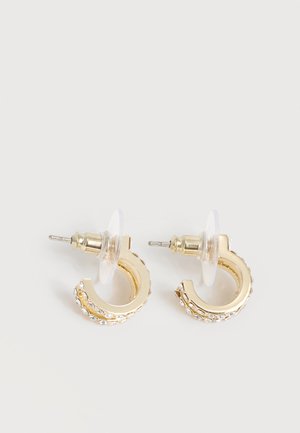 Μικρά χρυσά σκουλαρίκια με ενσωματωμένους διαφανείς στρας και διαφανείς πλάτες τοποθετημένα σε λευκή επιφάνεια.