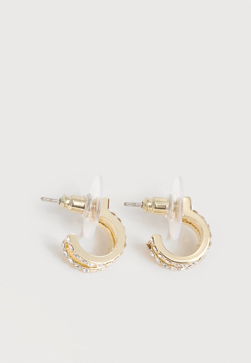 Μικρά χρυσά σκουλαρίκια με ενσωματωμένους διαφανείς στρας και διαφανείς πλάτες τοποθετημένα σε λευκή επιφάνεια.