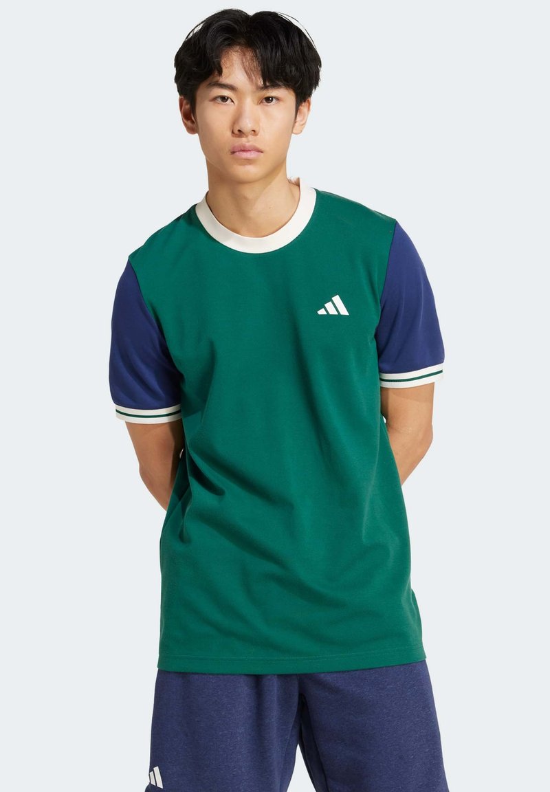 Green cotton t-shirt with blue short sleeves, white collar, and a small white logo on the left chest. Paired with navy shorts.