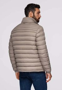 Lichte beige pufferjack met een hoge kraag, verticale gestikte secties en elastische manchetten; gedragen met donkere spijkerbroeken.