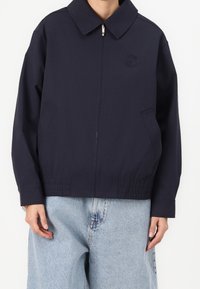 Donkerblauwe jas met een puntige kraag, ritssluiting aan de voorkant en twee zijzakken; voorzien van een elastische zoom en een lichte, gladde stoftextuur.