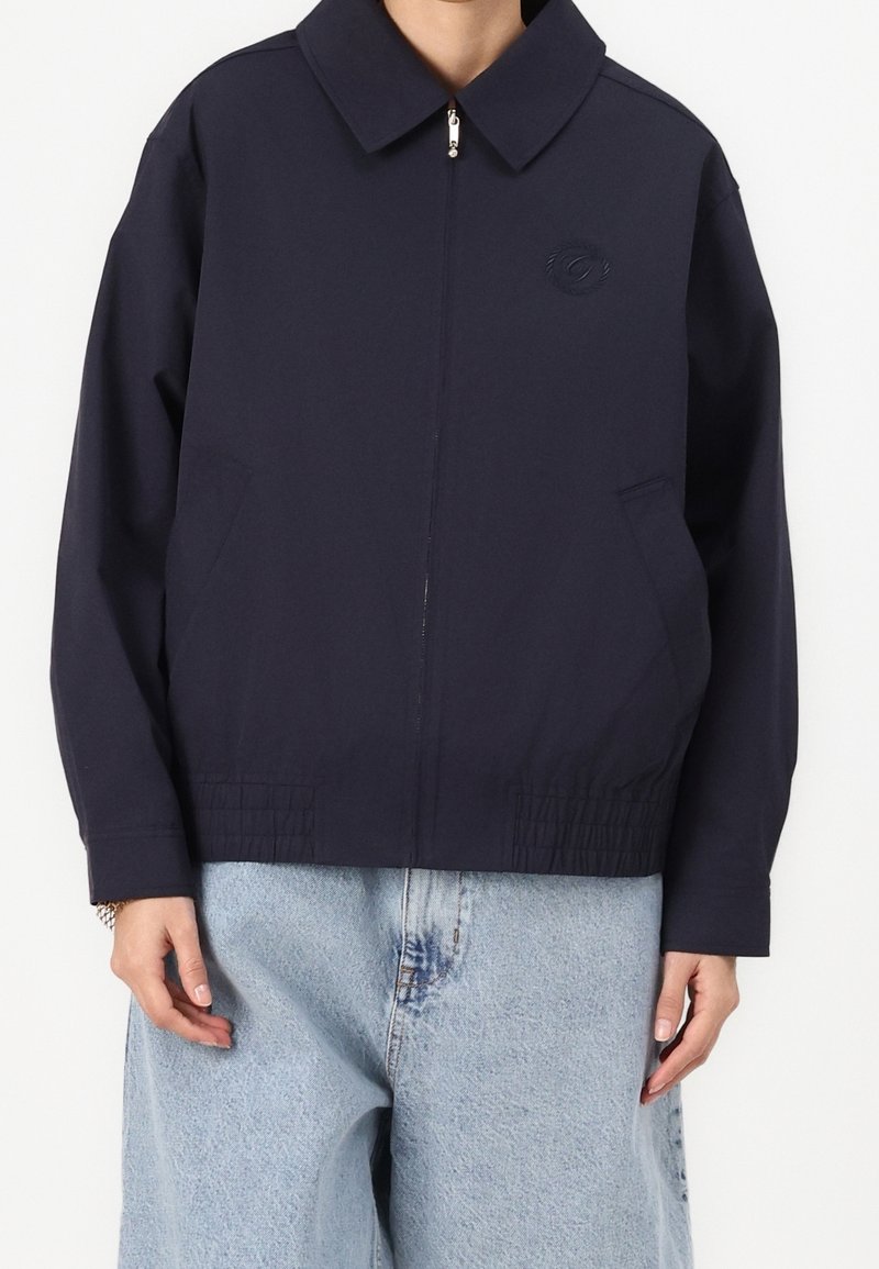 Donkerblauwe jas met een puntige kraag, ritssluiting aan de voorkant en twee zijzakken; voorzien van een elastische zoom en een lichte, gladde stoftextuur.