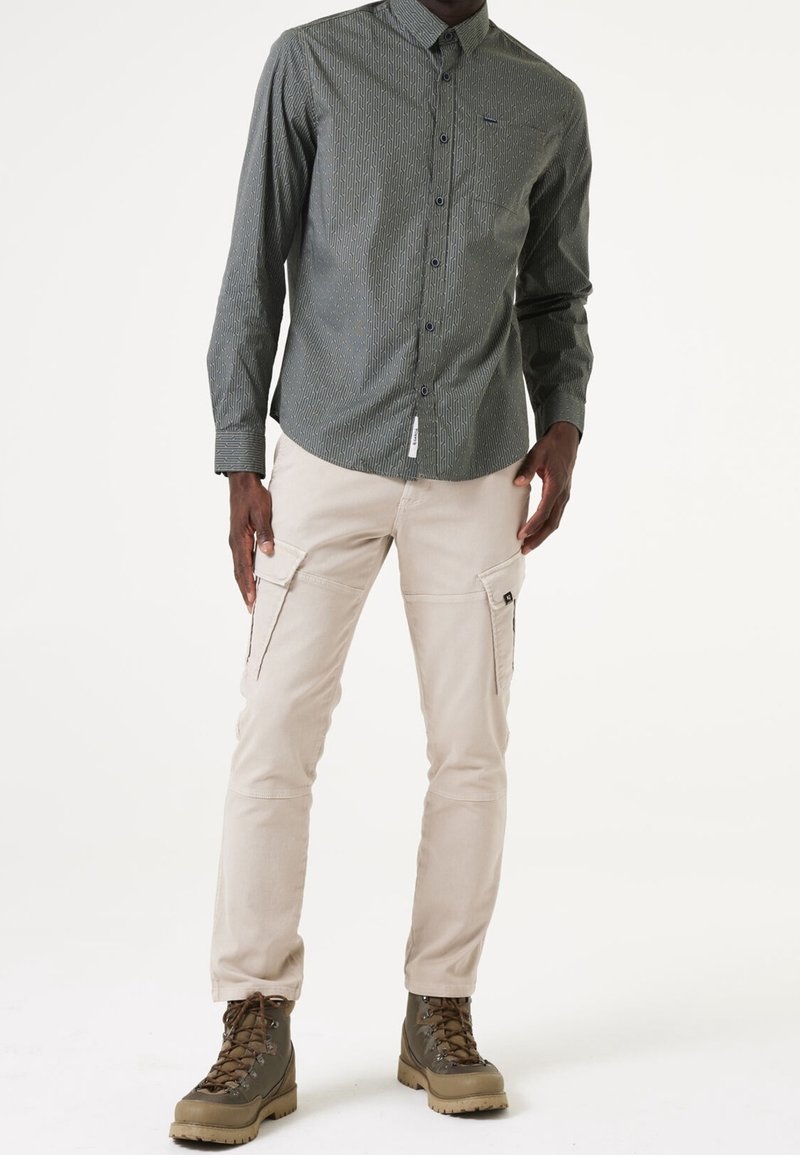 Homme portant une chemise grise à manches longues à motifs, un pantalon cargo beige clair avec poches latérales et des bottes marron à lacets, debout sur un fond blanc.