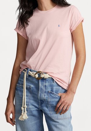 Frau trägt ein hell rosa T-Shirt mit kurzen Ärmeln, das in eine hoch taillierte blaue Jeans mit einem geknoteten Seilgürtel und großen Fronttaschen gesteckt ist.