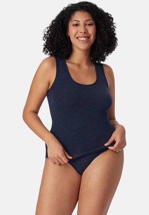 Donna con capelli ricci sorridente, che indossa una canottiera blu navy e biancheria intima abbinata, mentre sistema l'orlo inferiore della canottiera.