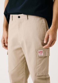 Beige cargobroeken hebben een zijzak met een rood-wit "Red Bull Racing" label. Gemaakt van duurzaam materiaal met een klassieke pasvorm en rechte pijpen.