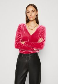 Langermet topp med V-hals i lys rosa fløyel, kombinert med høyt liv sorte skinnbukser. Har glatt tekstur og tettsittende design.
