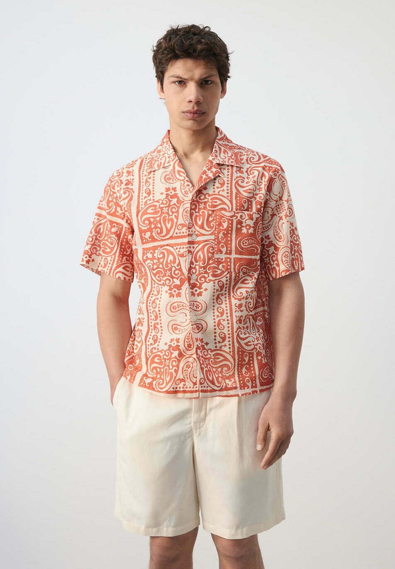Jeune homme portant une chemise à manches courtes à motifs orange et blanc et un short couleur crème, debout devant un fond uni.