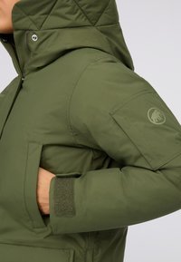 Giubbotto verde imbottito con cappuccio, caratterizzato da un design trapuntato, chiusura frontale con zip e polsini con chiusura in Velcro. Logo sulla manica sinistra.