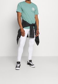 Une personne portant un t-shirt turquoise et un jean skinny blanc se tient avec une veste noire nouée autour de la taille, accompagnée de sneakers noires.