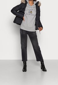 Giacca nera imbottita con colletto in pelliccia, felpa grigia con logo, jeans neri a vita alta e stivaletti neri con tacco. Texture lisce, design casual.