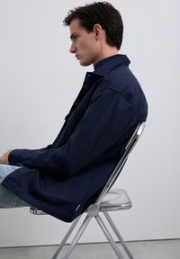 Joven con cabello oscuro, vestido con chaqueta navy y jeans de mezclilla claros, sentado de lado en una silla de metal contra una pared sencilla.