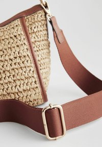 Sac en fibre naturelle tissé avec une sangle en tissu marron. Présente un détail en matériel rectangulaire doré. Texture lisse et forme structurée.