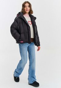 Czarna pikowana kurtka puchowa z kapturem, z poziomym pikowaniem, zapinana na zamek błyskawiczny i z prążkowanymi detalami, zestawiona z jasnoniebieskimi dżinsami i czarnymi butami.